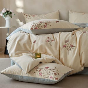 Pode incluir: Conjunto de roupa de cama creme com bordados florais. O conjunto inclui uma capa de edredão e fronhas, todas adornadas com delicadas flores rosa e borboletas. Um pequeno castiçal verde está na cama. As almofadas têm uma borda azul clara.