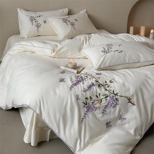 May include: Cream-colored bedding set featuring embroidered wisteria and butterfly designs. The set includes a duvet cover, pillows, and a fitted sheet. A small book and lit candle are placed on the bed, adding a touch of elegance to the bedroom decor.