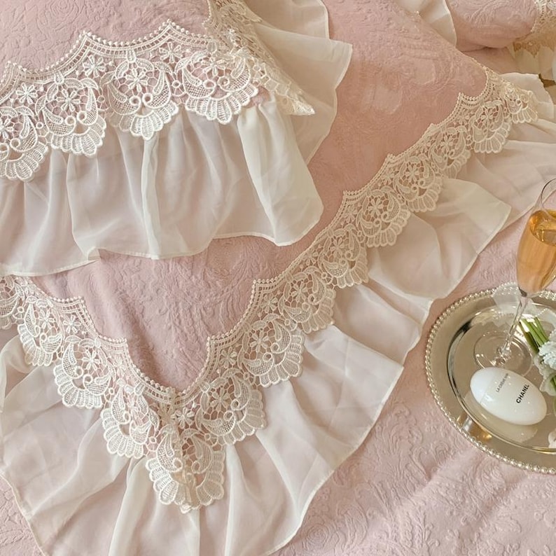 Pode incluir: Close-up de uma colcha rosa com acabamento em renda branca e babados. A renda apresenta intrincados desenhos florais. Uma bandeja prateada com uma ta&ccedil;a de champanhe e um pequeno frasco sugere um tema luxuoso.