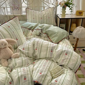 May include: A bed with a white metal headboard features a white and green striped bedding set. The set includes a duvet, pillows, and a comforter, all with cherry embroidery. A teddy bear and a pearl beaded bag are on the bed.