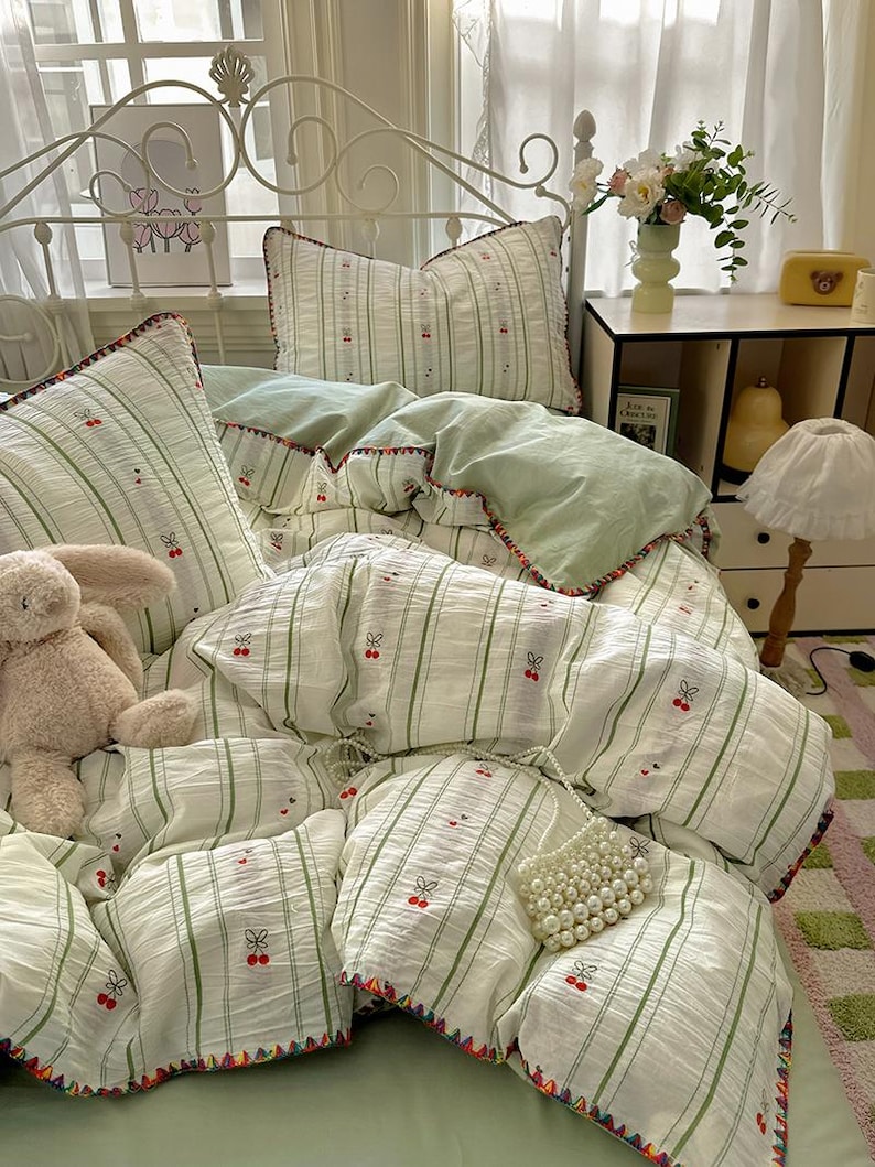 May include: A bed with a white metal headboard features a white and green striped bedding set. The set includes a duvet, pillows, and a comforter, all with cherry embroidery. A teddy bear and a pearl beaded bag are on the bed.