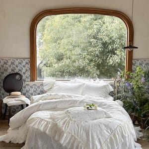 Pode incluir: Uma cama branca com uma saia de cama com babados e roupa de cama a condizer. A cama está em frente a uma grande janela com moldura de madeira. Uma cadeira pequena com um chapéu está à esquerda da cama. O quarto tem um ambiente acolhedor.