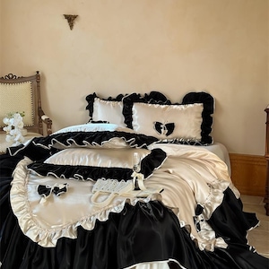 Puede incluir: Un lujoso juego de cama con una combinación de colores negro y crema. La ropa de cama incluye una falda de cama con volantes, almohadas decorativas con lazos y un edredón de satén. Un collar de perlas y una copa de champán están sobre la cama.