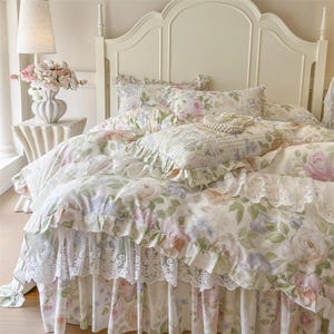 May include: A white bed with a floral patterned comforter, pillows, and bed skirt. The bedding features a delicate rose design in shades of pink, peach, and green. The bed has a white headboard and is in a well-lit room.