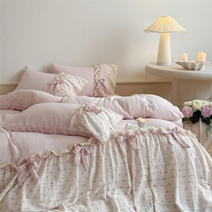 Pode incluir: Um conjunto de cama rosa com uma saia de cama estampada floral e almofadas a condizer. A roupa de cama tem bordas com babados e laços. Uma lâmpada branca e velas estão em uma mesa lateral. Um vaso de flores também é visível.