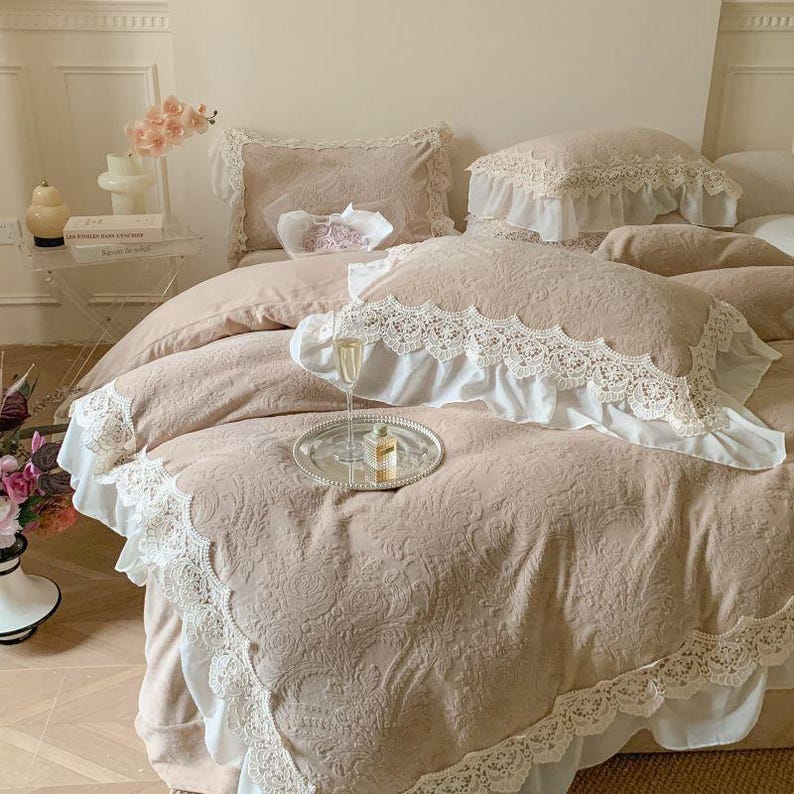 Pode incluir: Um conjunto de roupa de cama bege com intrincados padr&otilde;es florais e acabamento em renda branca. A roupa de cama inclui uma capa de edred&atilde;o, almofadas e uma almofada decorativa. Uma bandeja prateada com uma ta&ccedil;a de champanhe e um frasco de perfume est&aacute; na cama.