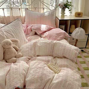 May include: A bed with a white metal headboard is covered in a pink and white striped bedding set. The set includes a duvet cover, pillows, and a pink fitted sheet. A stuffed bunny and a strand of pearls are on the bed.