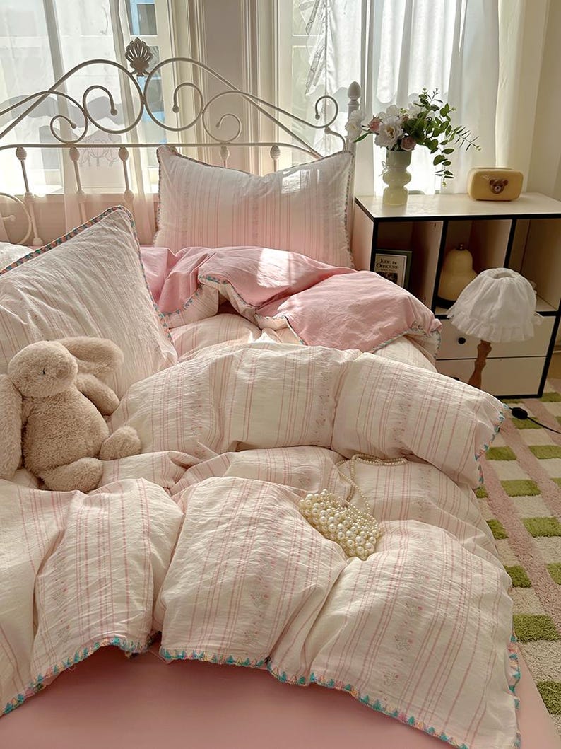 May include: A bed with a white metal headboard is dressed with a pink and white striped bedding set. The set includes a duvet cover, pillows, and a pink fitted sheet. A stuffed bunny and a string of pearls are on the bed.