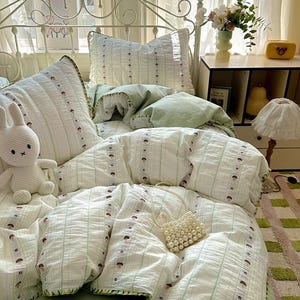 May include: A bed with a white metal frame is dressed with a light green and white bedding set. The set includes a duvet, pillows, and a fitted sheet. The duvet and pillows have a striped pattern with small brown mushroom designs. A white stuffed bunny and a pearl beaded purse are on the bed.
