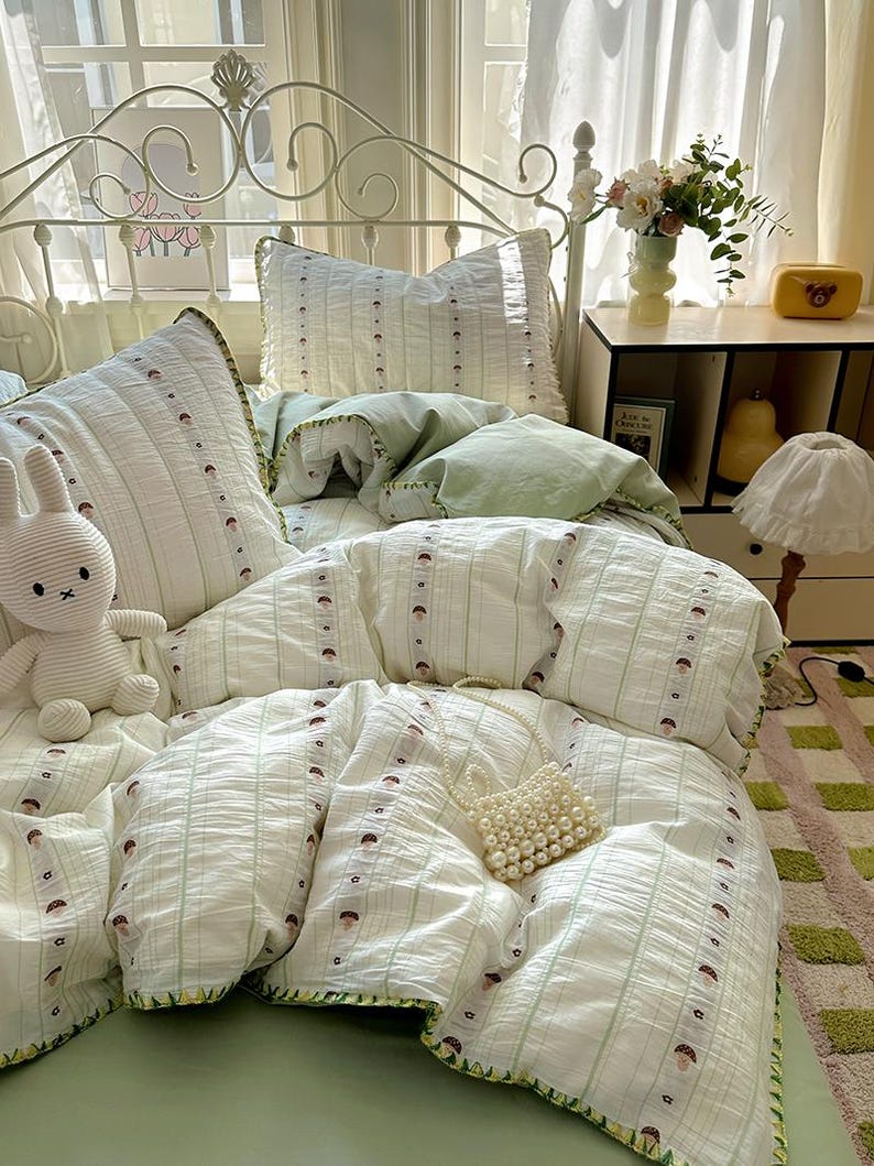 May include: A bed with a white metal frame is dressed with a light green and white bedding set. The set includes a duvet, pillows, and a fitted sheet. The duvet and pillows have a striped pattern with small brown mushroom designs. A white stuffed bunny and a pearl beaded purse are on the bed.