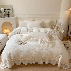 Pode incluir: Um conjunto de cama branco com um padr&atilde;o floral, com uma capa de edred&atilde;o, almofadas e uma saia de cama. O conjunto &eacute; adornado com renda delicada em um tom rosa suave, criando uma est&eacute;tica rom&acirc;ntica e elegante. Uma bandeja com guloseimas e uma ta&ccedil;a de champanhe est&atilde;o na cama.
