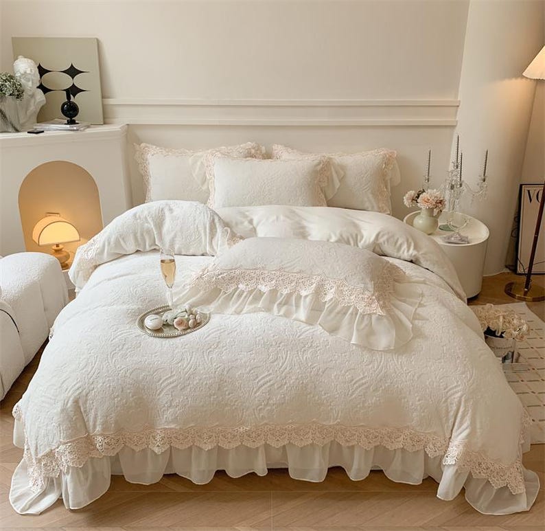 Pode incluir: Um conjunto de cama branco com um padr&atilde;o floral, com uma capa de edred&atilde;o, almofadas e uma saia de cama. O conjunto &eacute; adornado com renda delicada em um tom rosa suave, criando uma est&eacute;tica rom&acirc;ntica e elegante. Uma bandeja com guloseimas e uma ta&ccedil;a de champanhe est&atilde;o na cama.