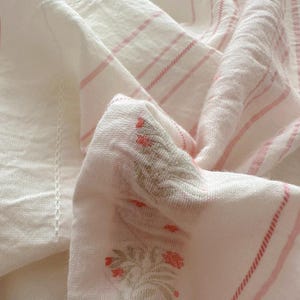 May include: A close-up shot of a white and pink striped fabric with floral embroidery. The fabric has a soft texture, possibly linen or cotton. The pink stripes are delicate. The floral embroidery features red flowers.