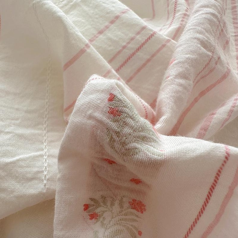 May include: A close-up shot of a white and pink striped fabric with floral embroidery. The fabric has a soft texture, possibly linen or cotton. The pink stripes are delicate. The floral embroidery features red flowers.