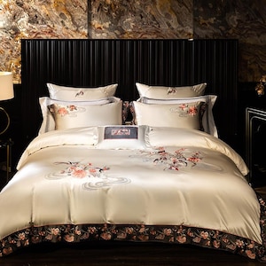 May include: Cream-coloured bedding set with floral embroidery in pink, red, and grey. The set includes a duvet cover, pillowcases, and decorative cushions. The duvet cover and pillowcases feature a floral border. The headboard is black.