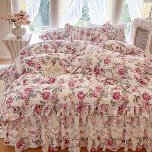 Op de afbeelding: Bloemen beddengoedset met een dekbedovertrek, kussenslopen en een bedrok. De set heeft een crèmekleurige achtergrond met roze en rode rozen en groene bladeren. De bedrok heeft een kanten rand.