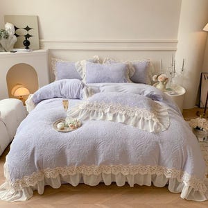 Pode incluir: Um conjunto de roupa de cama lil&aacute;s claro com padr&atilde;o floral e acabamento em renda branca. O conjunto inclui edred&atilde;o, almofadas e uma saia de cama. Uma bandeja decorativa com flores e uma ta&ccedil;a de champanhe est&aacute; na cama. Tema rom&acirc;ntico.
