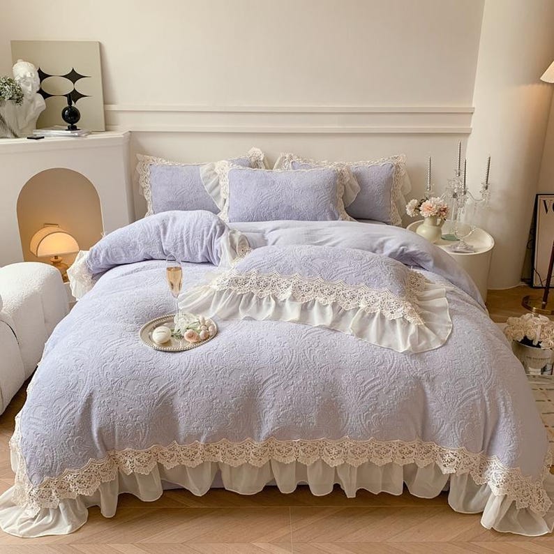 Pode incluir: Um conjunto de roupa de cama lil&aacute;s claro com padr&atilde;o floral e acabamento em renda branca. O conjunto inclui edred&atilde;o, almofadas e uma saia de cama. Uma bandeja decorativa com flores e uma ta&ccedil;a de champanhe est&aacute; na cama. Tema rom&acirc;ntico.
