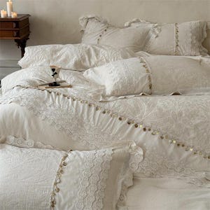 May include: Cream-colored bedding set featuring a duvet cover and several pillows. The duvet and pillowcases are adorned with intricate lace trim and small, gold-colored embellishments. The bedding has a floral pattern.