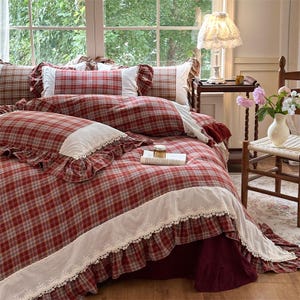 Könnte beinhalten: Ein rot-weiß kariertes Bettwäsche-Set mit Rüschen und weißer Spitzenborte. Das Set umfasst einen Bettbezug, Kissenbezüge und einen Bettrock. Ein Buch und ein kleines Glas liegen auf dem Bett. Die Szene wird von einer Lampe beleuchtet.