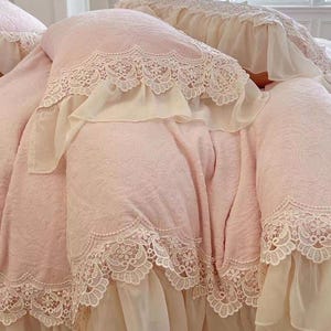 Pode incluir: Um edredom rosa claro com um padr&atilde;o floral e acabamento em renda branca. O edredom tem uma borda com babados e &eacute; feito de um tecido macio e texturizado. A renda adiciona um toque delicado &agrave; roupa de cama.