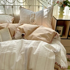 May include: A bed with a white metal frame and layered bedding. The bedding includes a cream-coloured duvet cover and striped sheets with decorative trim. A small, white stuffed bunny sits on the pillows. A book and a vase of flowers are on a nearby bedside table.