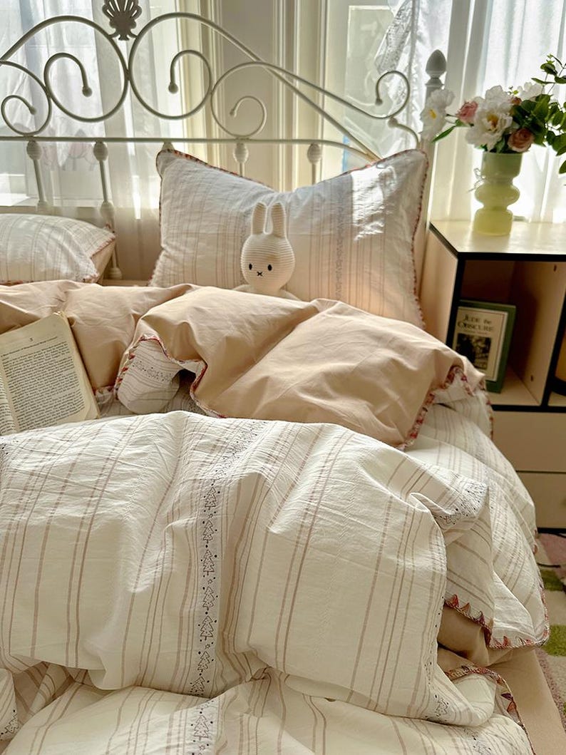 May include: A bed with a white metal frame and layered bedding. The bedding includes a cream-coloured duvet cover and striped sheets with decorative trim. A small, white stuffed bunny sits on the pillows. A book and a vase of flowers are on a nearby bedside table.