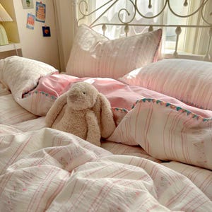 May include: A bed with pink and white striped bedding, a plush bunny, and pillows. The duvet and pillows have a delicate floral pattern. The overall aesthetic is soft and inviting, with a focus on comfort and relaxation.