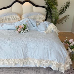 Pode incluir: Um edredom azul claro com um padrão floral em relevo e uma borda de renda creme. A cama é feita com fronhas azuis claras combinando com acabamento em renda creme. Um buquê de flores rosa e brancas repousa na cama.