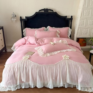May include: A pink bedding set featuring a duvet cover and pillowcases with white ruffled trim and decorative fabric flower accents. The set includes a rectangular bolster pillow and a standard pillow, all in a solid pink color with white ruffles.
