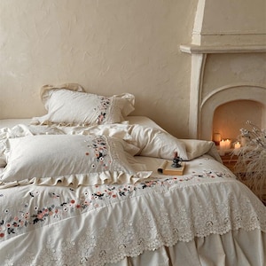 Puede incluir: Juego de cama color crema con bordados florales y ribete de encaje. El juego incluye una funda nórdica, fundas de almohada y una falda de cama. El bordado presenta flores rosas, rojas y verdes. La cama está contra una pared de tonos neutros.