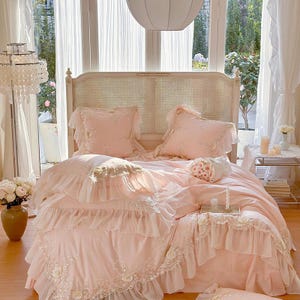 May include: A light pink bed set with ruffled edges and embroidered details. The set includes a comforter, pillows, and a bed skirt. The headboard is a light wood color with a woven design. The room has sheer white curtains and a chandelier.