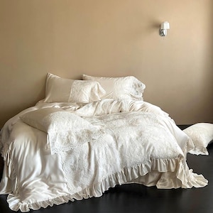 May include: Cream-coloured bedding set with a satin duvet cover and pillowcases, all with delicate lace trim. The bed has a ruffled bed skirt. The room has a neutral colour palette.