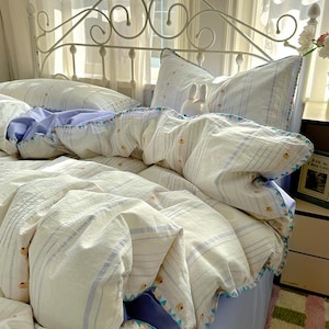 May include: A bed with a white metal headboard and bedding. The bedding features a duvet cover and pillows with a striped pattern in white and light blue. The duvet cover has a scalloped edge with a colourful trim.