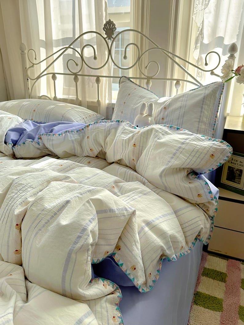 May include: A bed with a white metal headboard and bedding. The bedding features a duvet cover and pillows with a striped pattern in white and light blue. The duvet cover has a scalloped edge with a colourful trim.