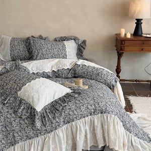 May include: A grey floral bedding set with white lace detailing. The set includes a duvet cover, pillowcases, and a decorative cushion. The bed is made up with a matching floral pattern and white lace trim. A wooden bedside table with a lamp and candles is visible.