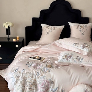 May include: A luxurious pink bedding set with embroidered floral designs. The set includes a duvet cover, pillows, and shams. The bed has a black velvet headboard and a black nightstand with a vase of white roses.