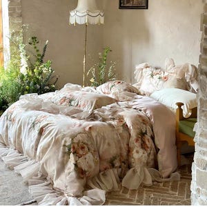 May include: A bed dressed with a floral duvet cover and pillowcases. The bedding has a cream base with pink and peach roses. The bed has a ruffled skirt. The room has a vintage aesthetic, with a floor lamp and brickwork.