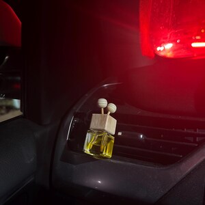 Pu&ograve; includere: Un piccolo profumatore per auto in vetro trasparente con una parte superiore in legno e due accenti sferici. Il liquido all'interno &egrave; giallo. Il profumatore &egrave; posto su una bocchetta di ventilazione del cruscotto dell'auto.