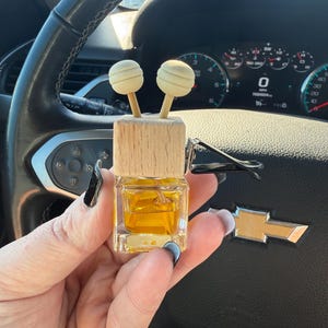 Pu&ograve; includere: Un profumatore per auto con una parte superiore in legno e due accenti sferici. La bottiglia di vetro trasparente contiene un liquido dorato. Il profumatore &egrave; tenuto davanti al volante e al cruscotto di un'auto.