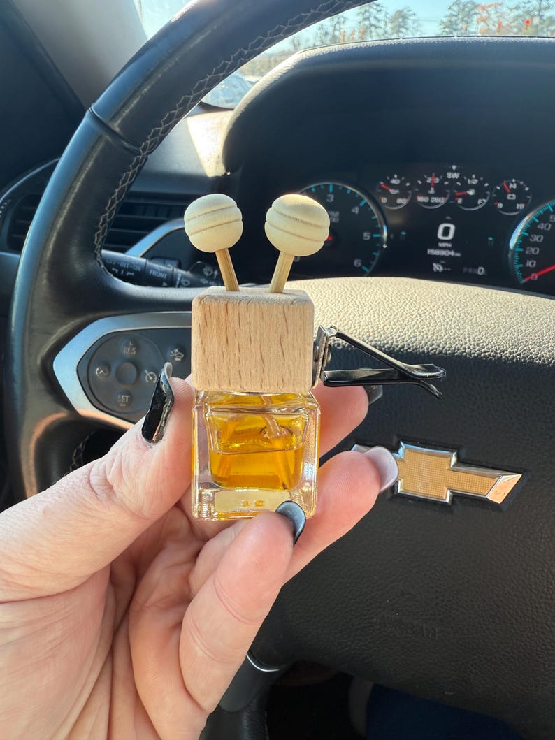 Pu&ograve; includere: Un profumatore per auto con una parte superiore in legno e due accenti sferici. La bottiglia di vetro trasparente contiene un liquido dorato. Il profumatore &egrave; tenuto davanti al volante e al cruscotto di un'auto.