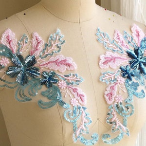 May include: Two blue and pink floral appliques with sequins on a white background. The appliques are made of lace and have a delicate, feminine design.