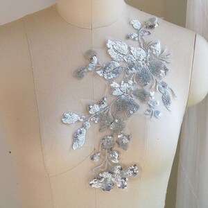 May include: Silver floral applique with sequins on a white mesh fabric. The applique features a cluster of flowers and leaves.