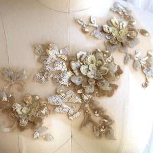 Op de afbeelding: Gouden en zilveren bloemenapplicatie met pailletten. De applicatie is op een witte stoffen achtergrond.