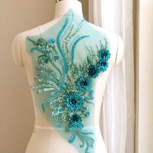 以下が含まれることがあります： 透き通った生地の背景に、ターコイズブルーとグリーンのフラワーモチーフのアップリケ。アップリケは涙のしずく型で、3Dの花の集まりが特徴です。