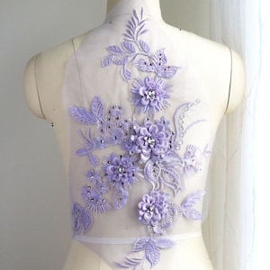 May include: A lavender floral applique with beaded accents. The applique is on a sheer mesh fabric and features a variety of flower shapes and leaves.