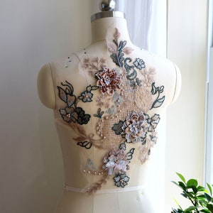 Peut inclure: Une applique en dentelle beige avec des motifs floraux et des accents de perles. L'applique présente une grande fleur multi-couches avec un centre en cuivre et des fleurs plus petites dans des tons de rose et de blanc.