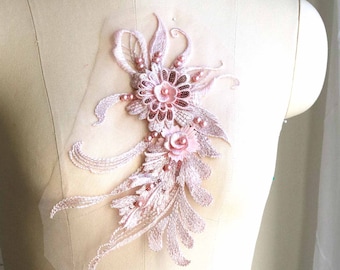 May include: A delicate, light pink floral applique with intricate bead and sequin detailing. The design features a central flower, smaller blossoms, and flowing leaf-like elements, all on a sheer mesh backing. This embellishment is ideal for adding a touch of elegance to garments.