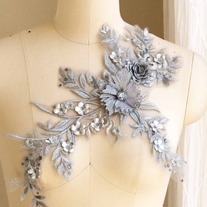 May include: Silver floral applique with sequins and beads on a white fabric background.