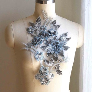 Może przedstawiać: Srebrna i niebieska aplikacja kwiatowa z cekinami i koralikami. Aplikacja znajduje się na białym manekinie.